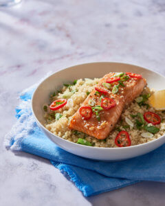 Honey-glazed salmon fillet on couscous with lemon and chili slices, on a blue napkin and marble surface.