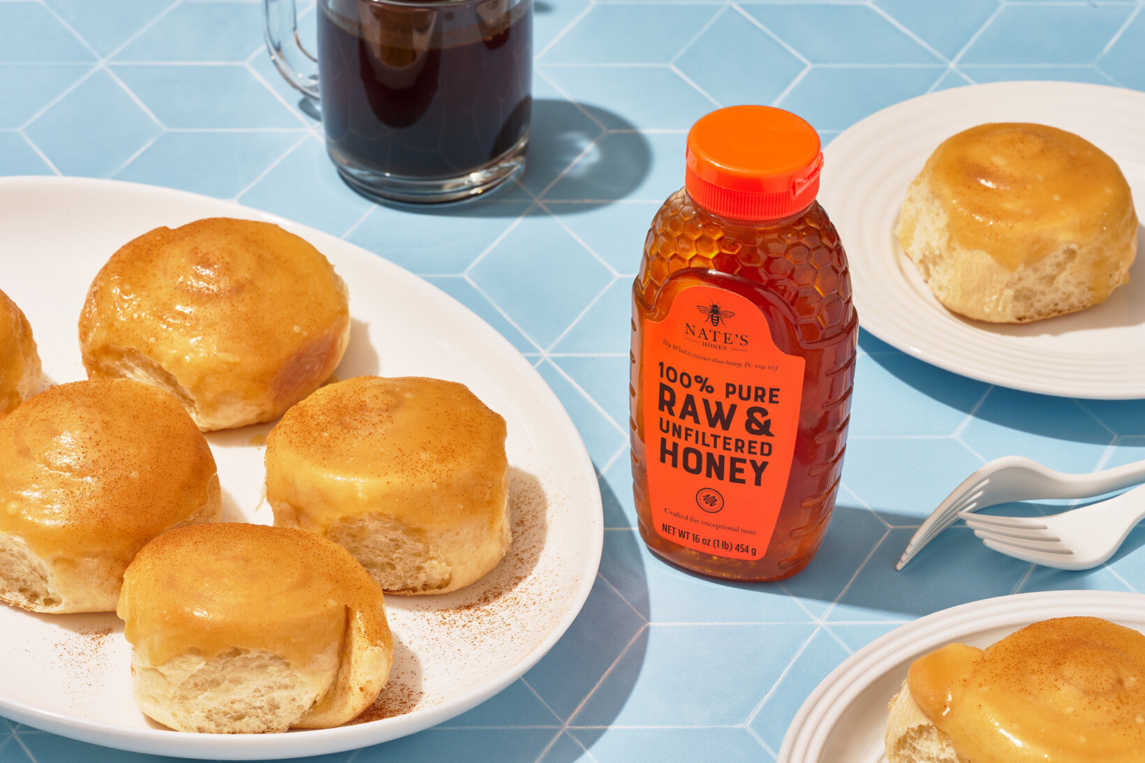 Golden honey buns on plates, with a bottle of Nate’s Honey, two forks and coffee.