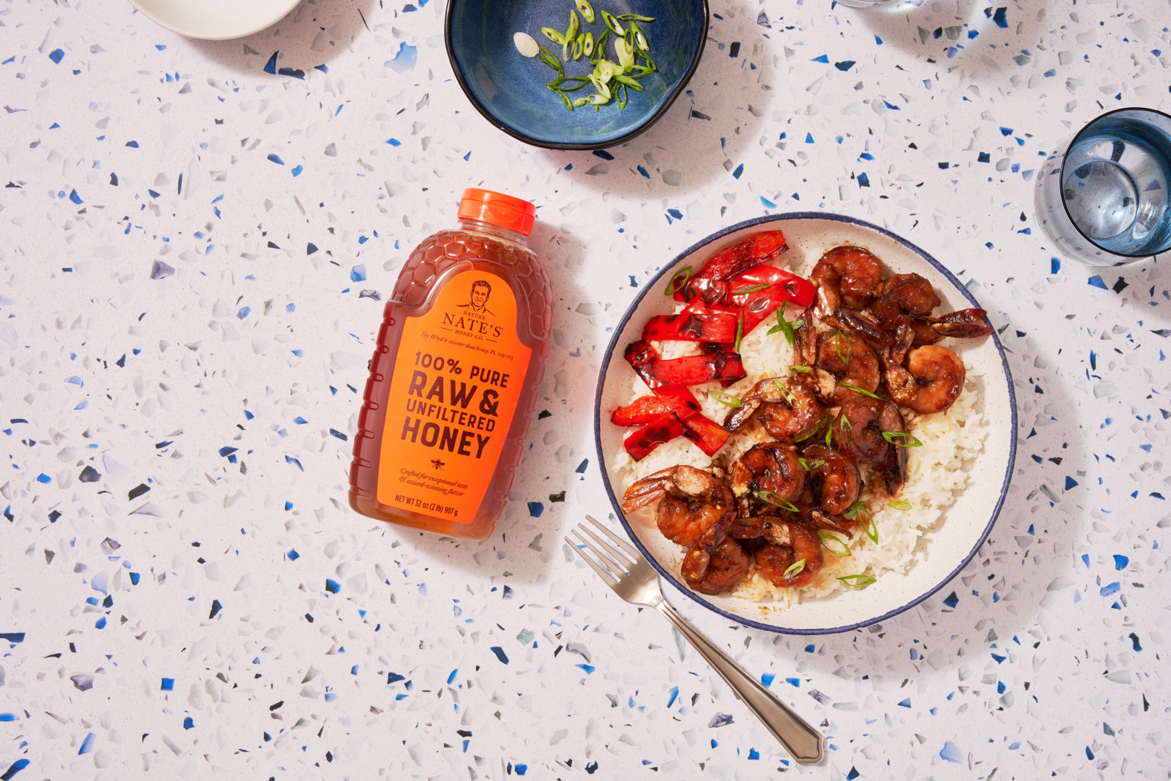 Honey-glazed shrimp, red peppers and rice on a plate, a bottle of Nate’s honey on a speckled counter, a fork and drink.