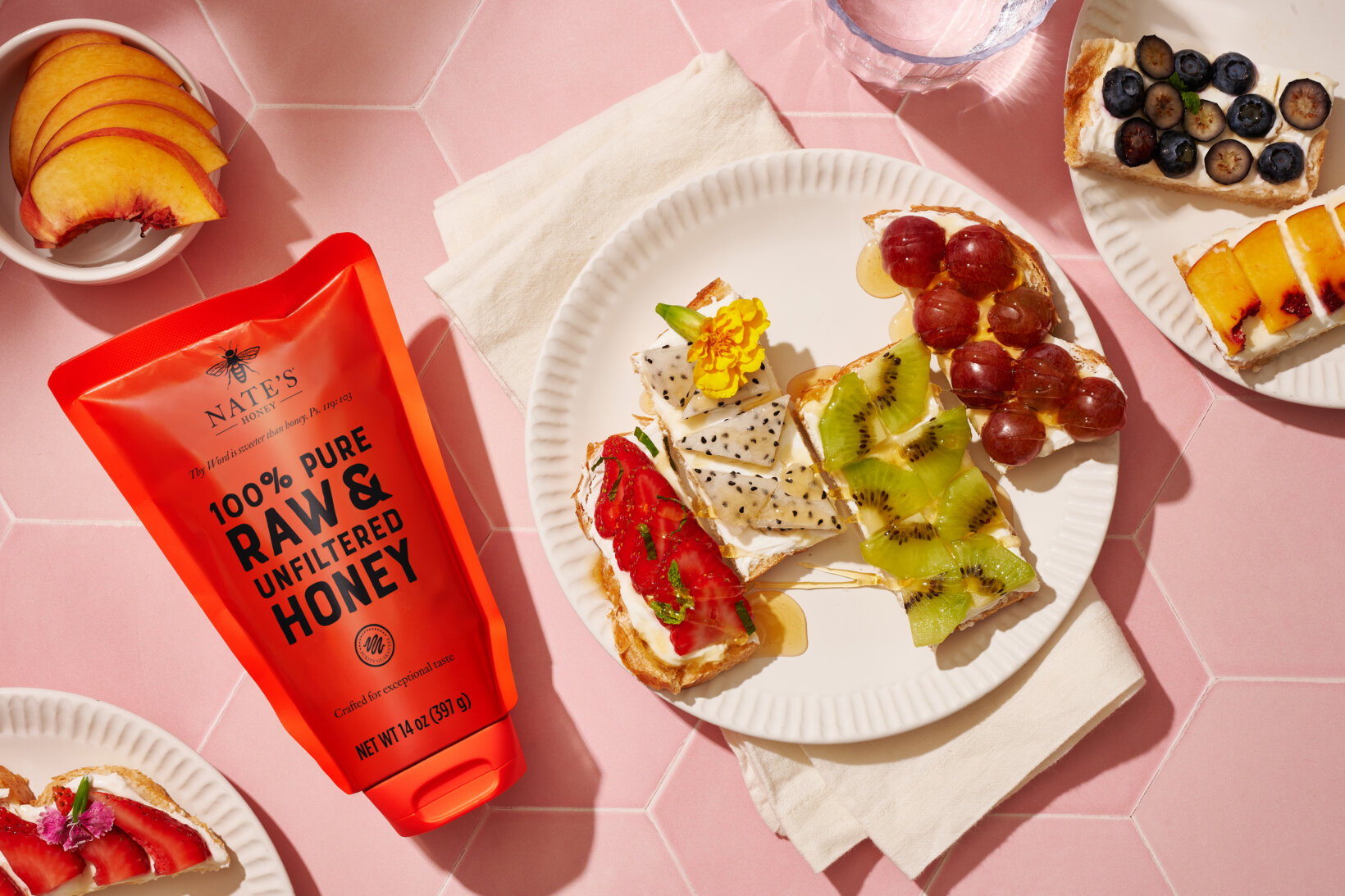 Open-faced fruit toasts on plates with a pouch of Nate’s Honey, against a pink background.