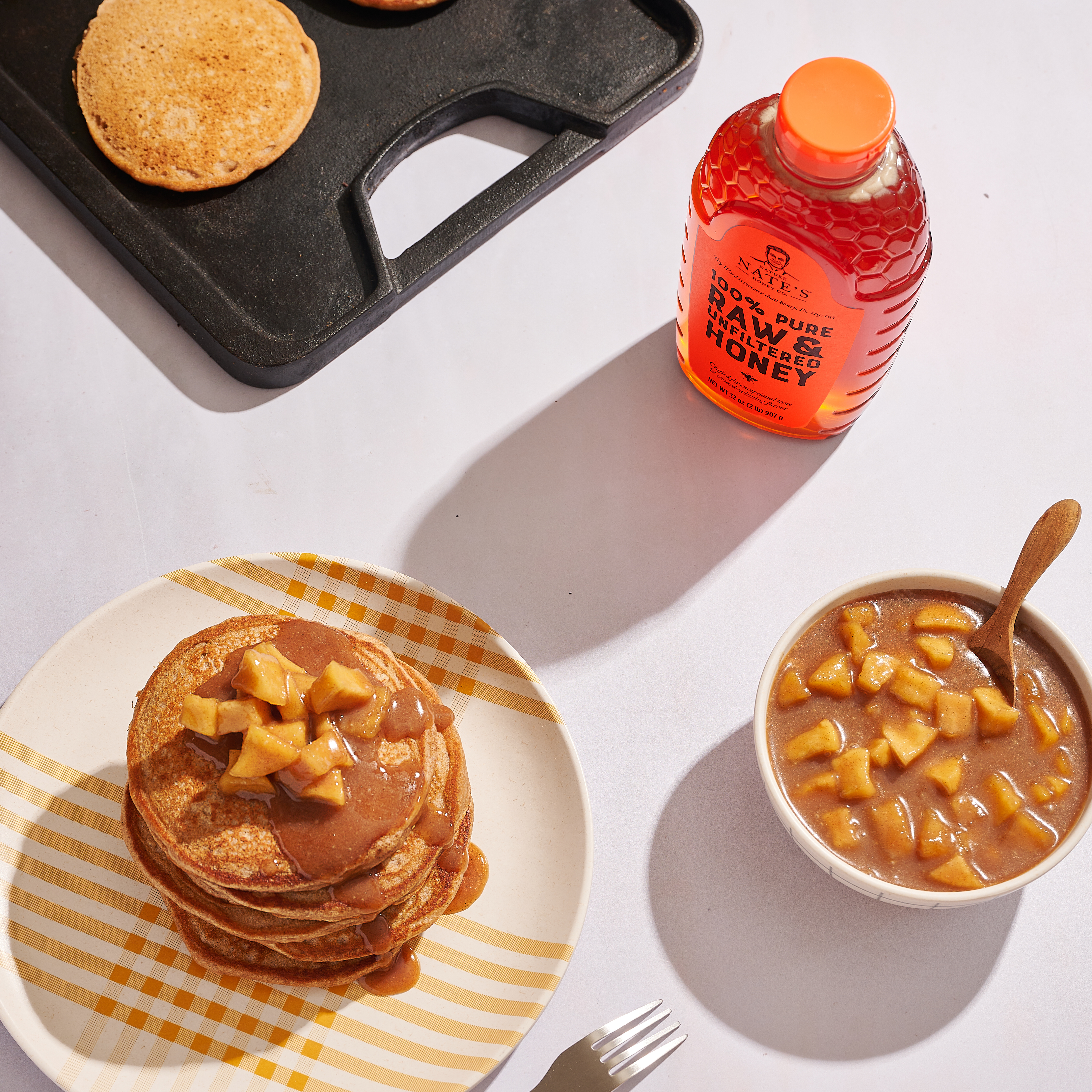 Plate of pancakes with apple topping next to a griddle and bottle of Nate's classic honey.
