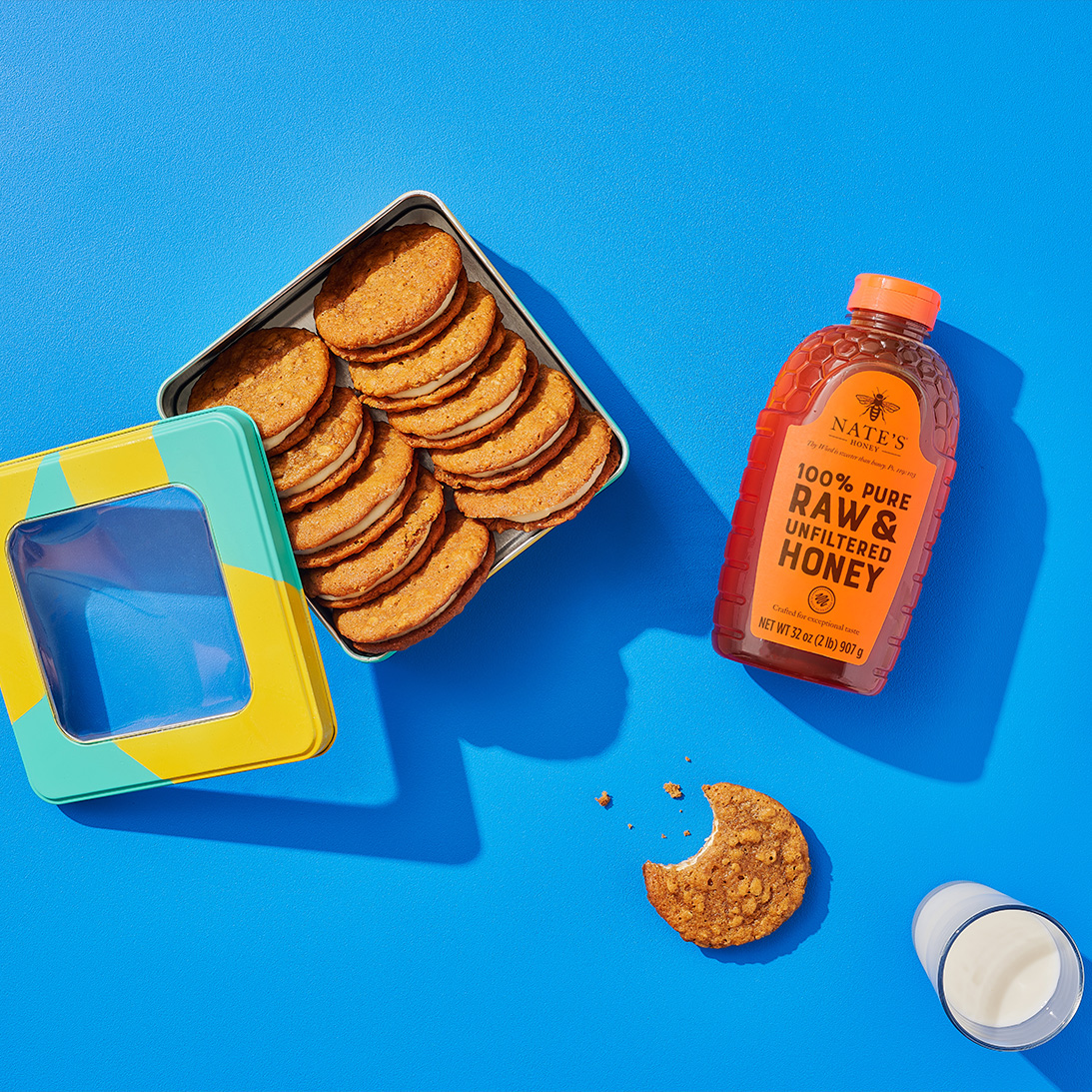 A tin of oatmeal crème pies next to a bottle of Nate’s honey, an oatmeal pie missing a bite and two glasses of milk.