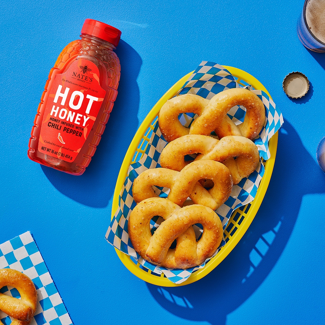 Nate’s hot honey bottle next to a basket of honeyed-pretzels on a blue background.
