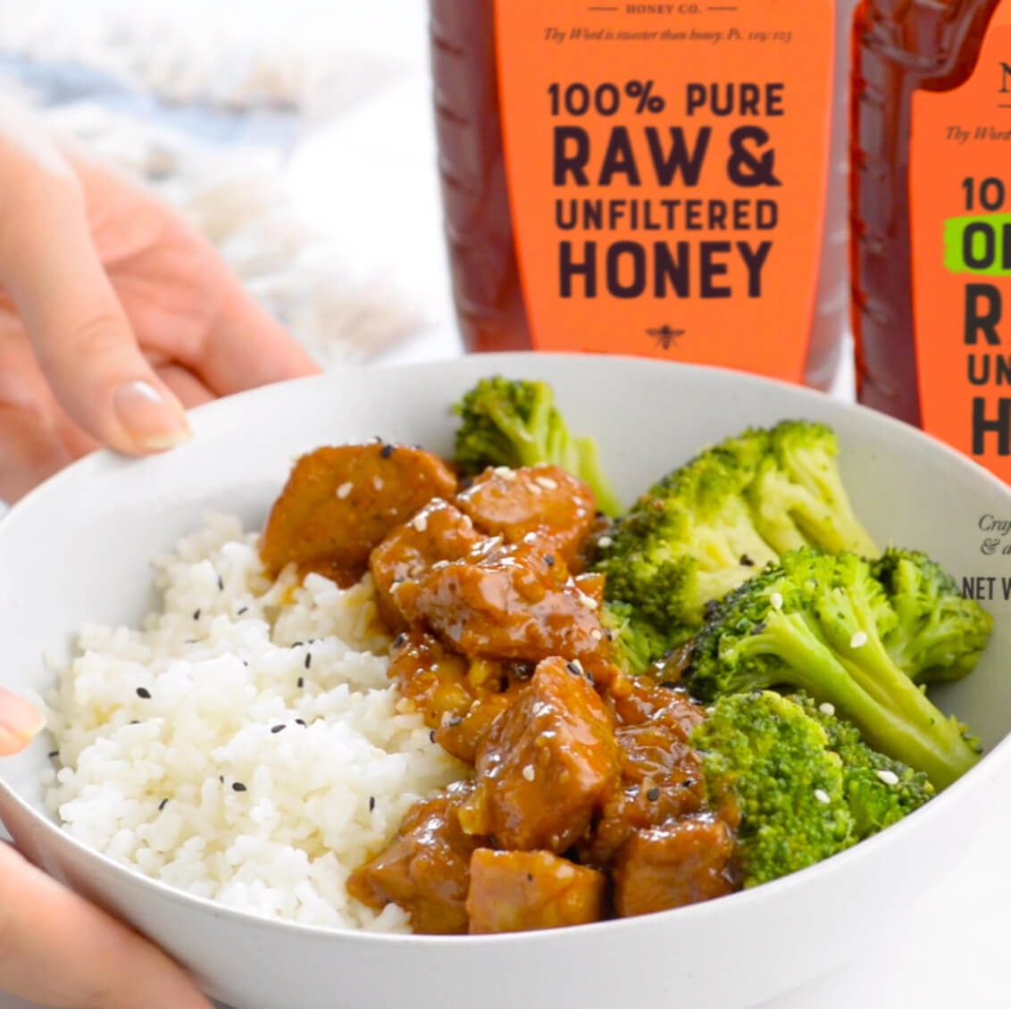 Hand holding bowl of rice with broccoli and glazed pork and Nate’s honey bottles in background.
