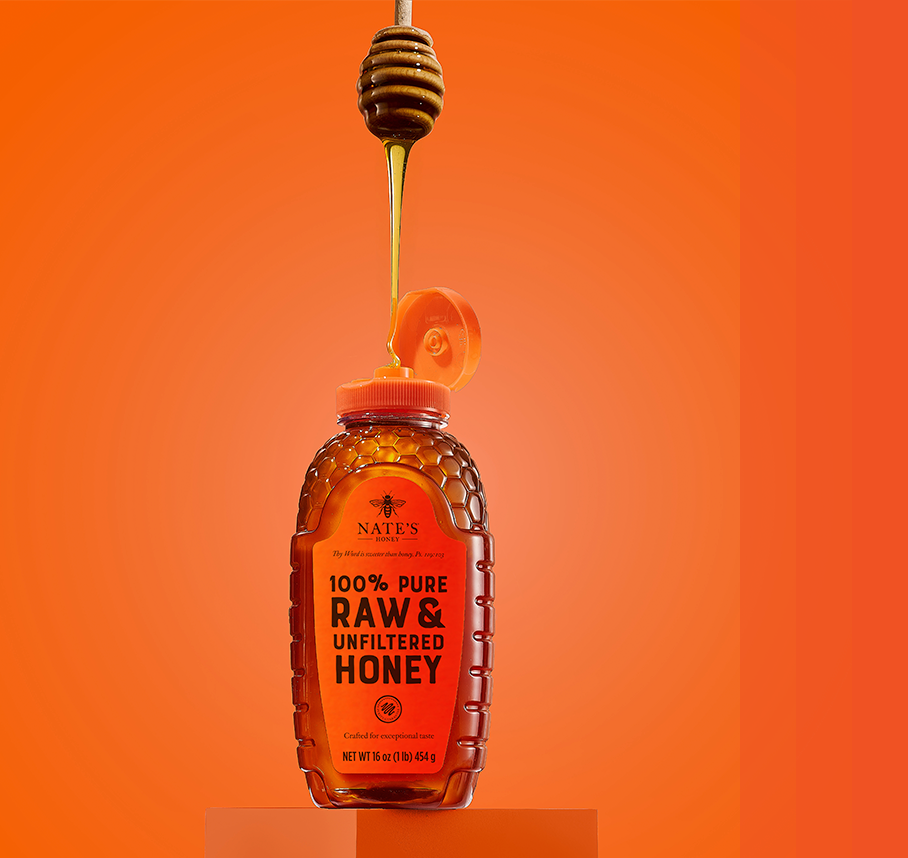 A bottle of Nate’s 100% Pure, Raw & Unfiltered Honey with the cap open, as honey drizzles from a dipper above the bottle against an orange background.