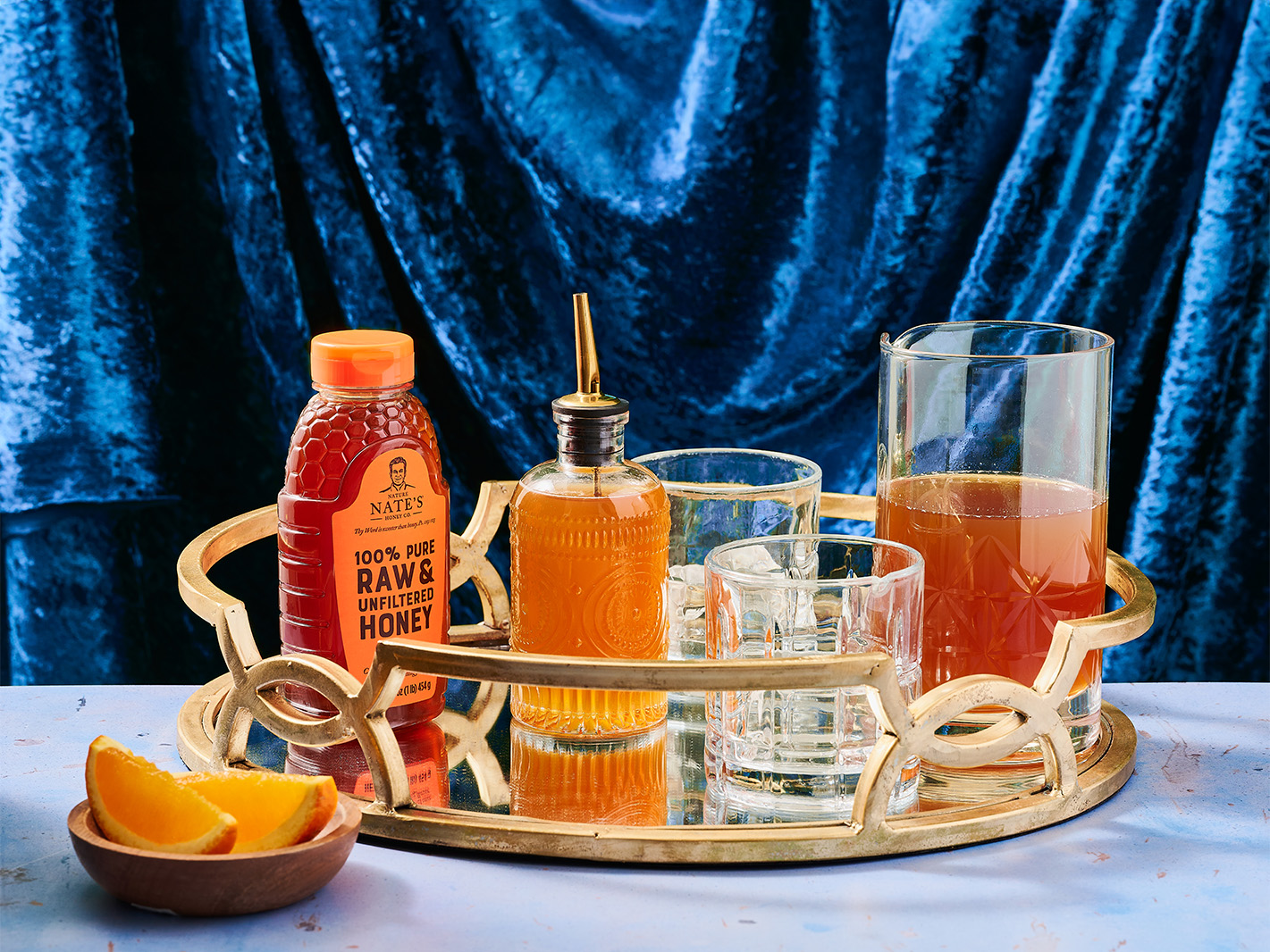 Nate's Honey with Holiday Drinks and Drinkware on golden tray with blue velvet in the background and orange slices in foreground.