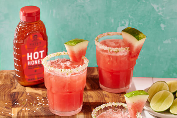 Two glasses of watermelon cocktail with salted rims, lime, watermelon slices and a bottle of Nate’s hot honey.