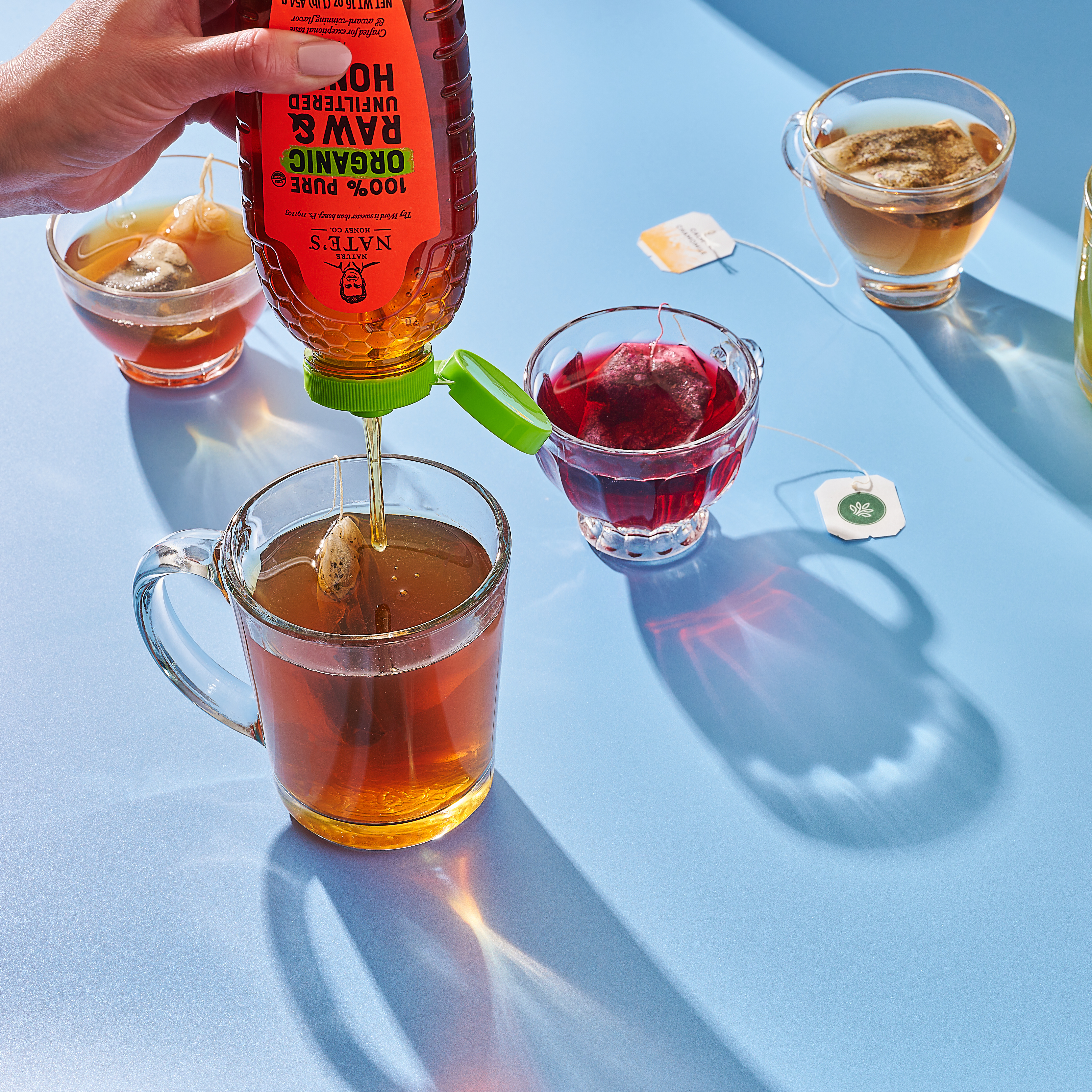 Clear mugs of assorted teas with a hand drizzling honey from a Nate's organic bottle.