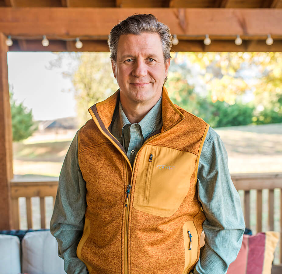 man in a vest and tie standing on a porch with a smile.