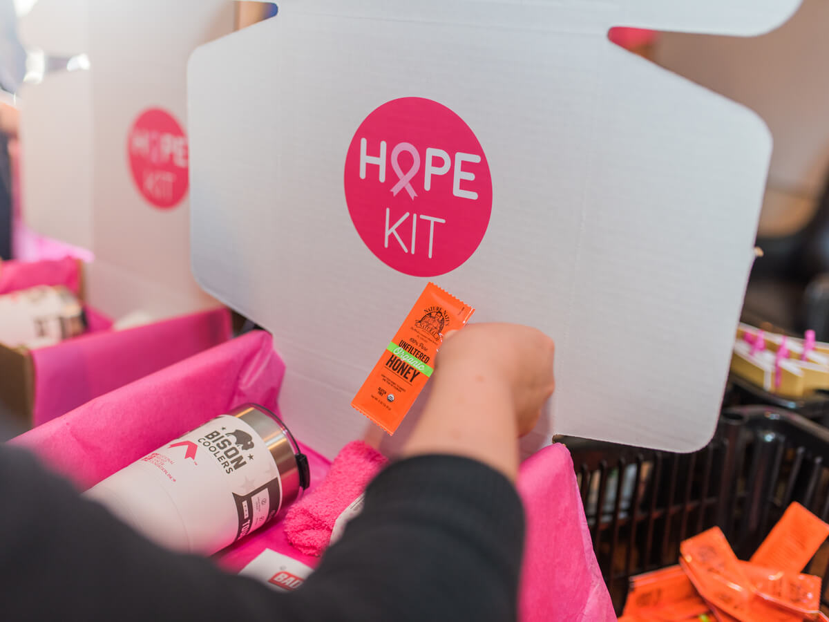 A hand assembling a HOPE KIT, box with a Nate’s honey mini and other items, with a pink interior.