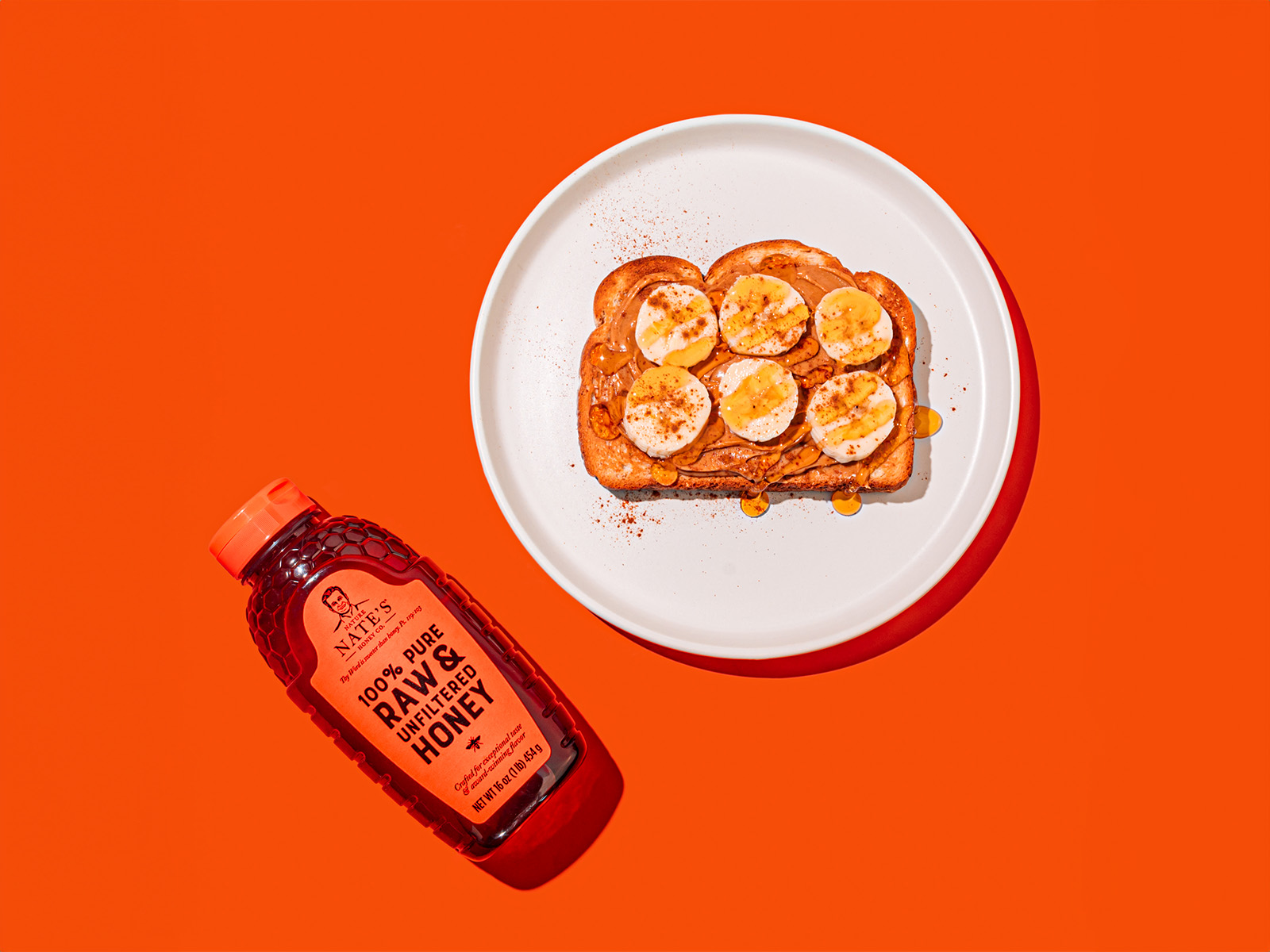 Peanut butter banana toast drizzled with honey on a white plate with a Nate’s honey bottle on orange background.