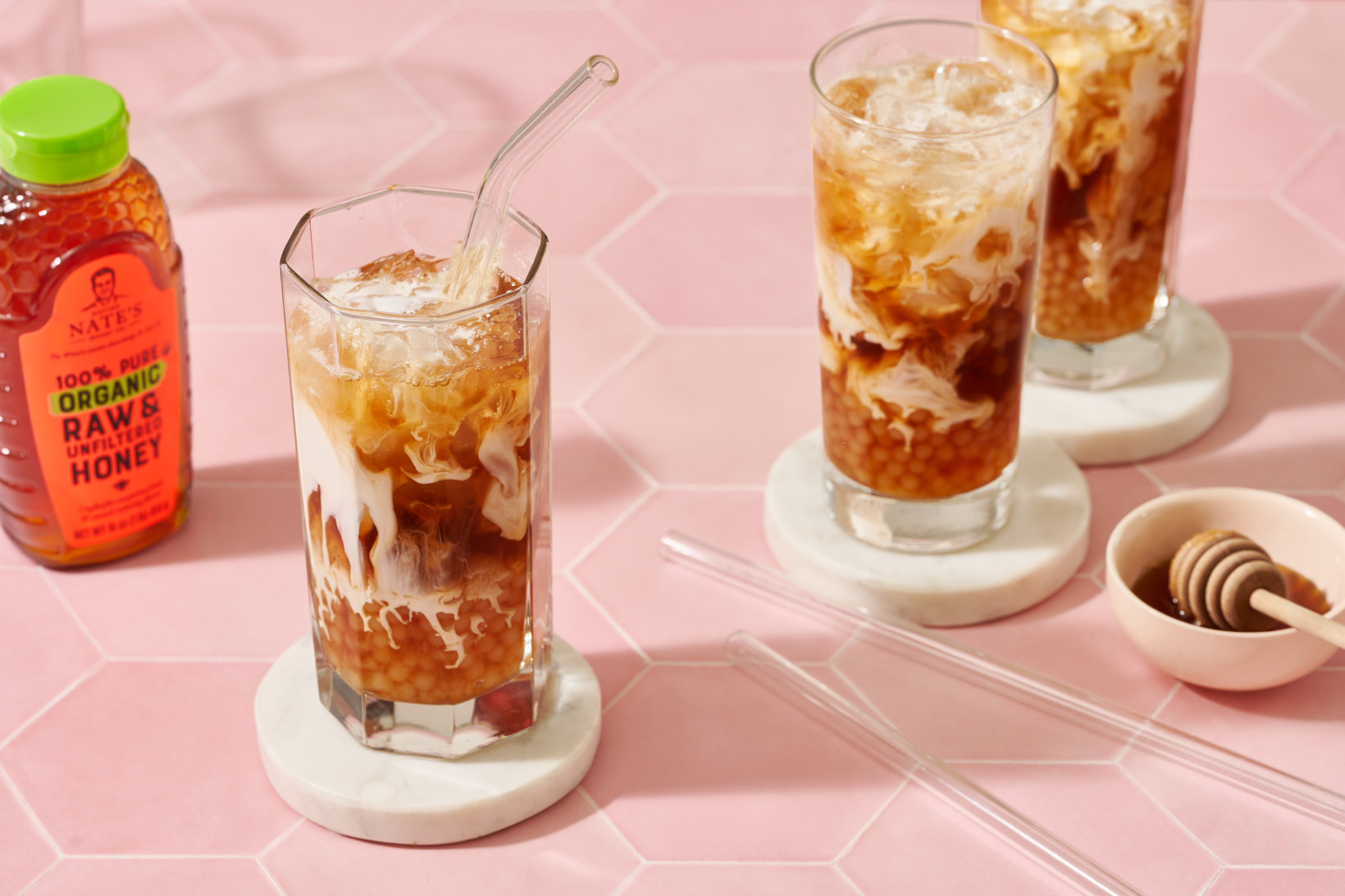 Tall glasses of iced coffee with cream on a pink surface next to a bottle of Nate’s organic honey and a honey dipper.
