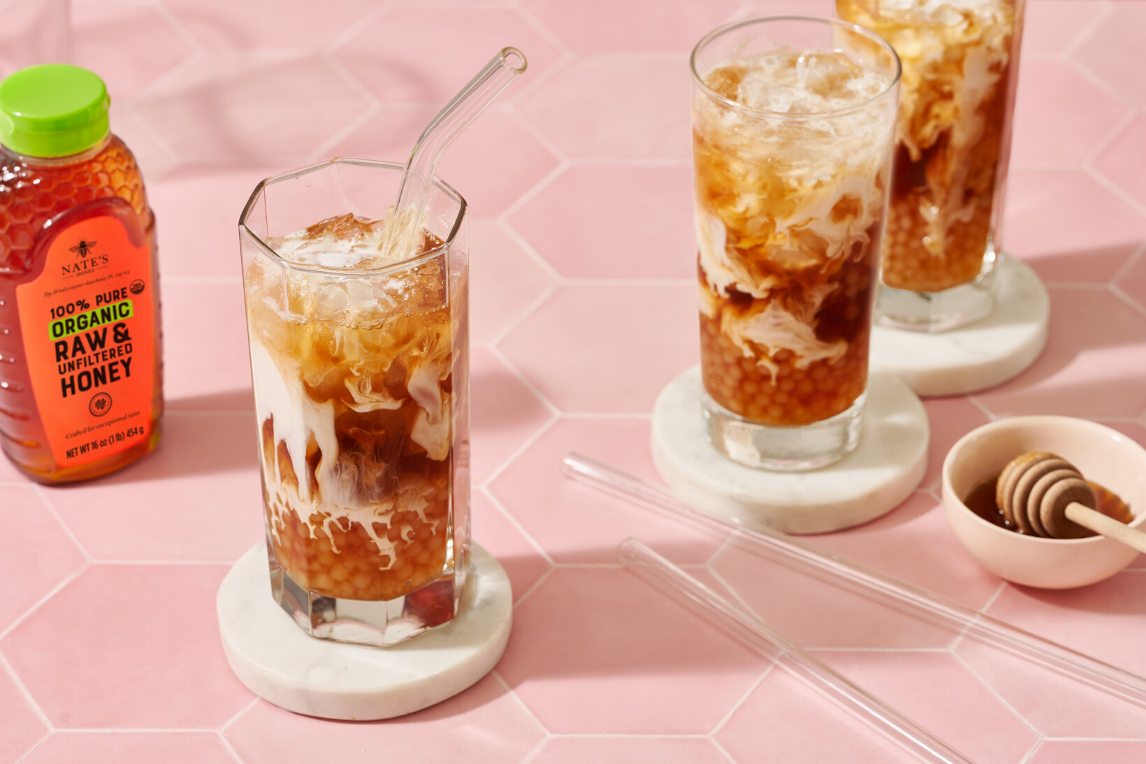 Tall glasses of iced coffee with cream on a pink surface next to a bottle of Nate’s organic honey and a honey dipper.