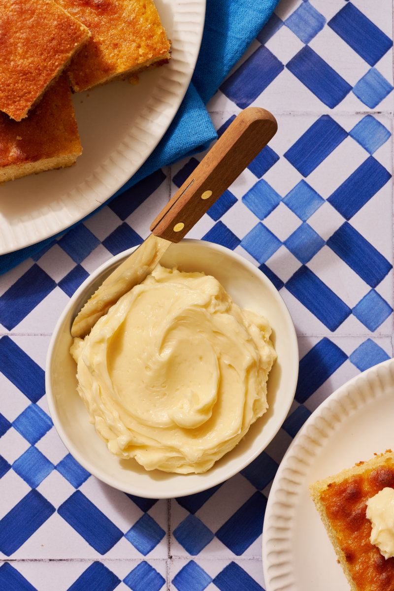 A bowl of creamy honey butter with a knife, toast with butter, all on a blue and white checkered tablecloth.