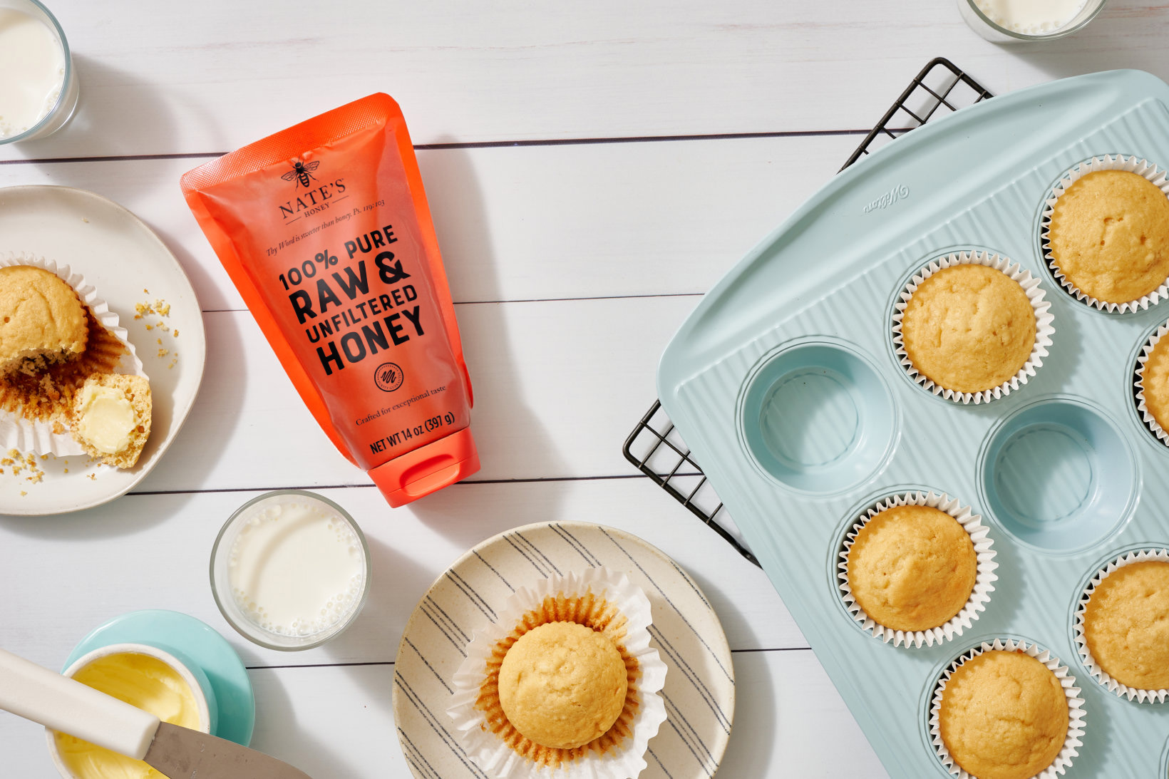 Honey muffins on a cooling rack and plates, with a Nate’s honey pouch, butter and milk on a white wooden table.