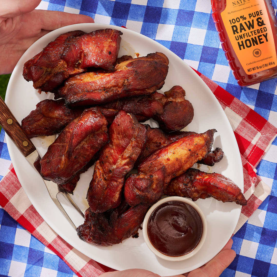 Hands serving a plate of BBQ pork ribs with a side of sauce and a Nate’s Honey bottle, on a blue and white blanket.