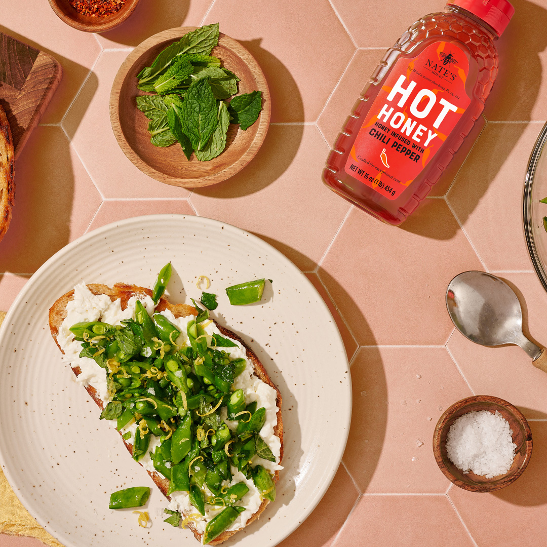 Burrata and snap pea toast on a plate, a bowl of snap peas, mint leaves and a bottle of Nate's Hot Honey on a pink table.