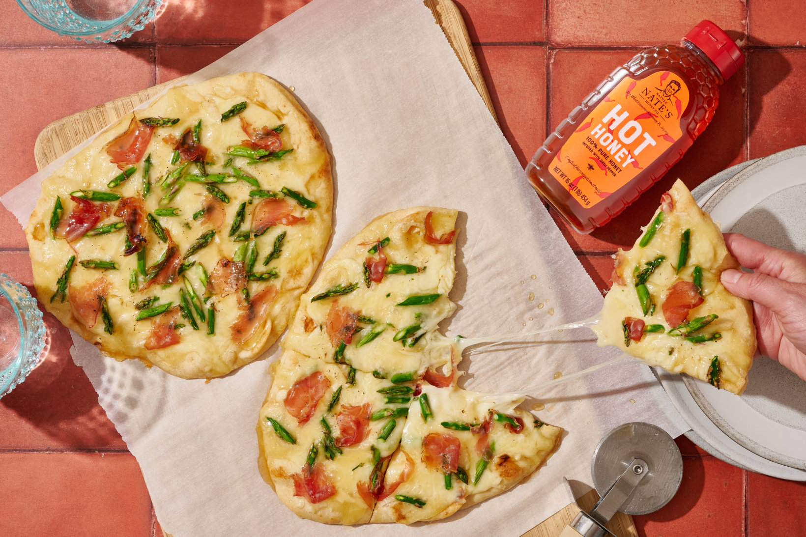 Two flatbread pizzas with prosciutto and asparagus and a bottle of Nate’s hot honey on a red tiled table.