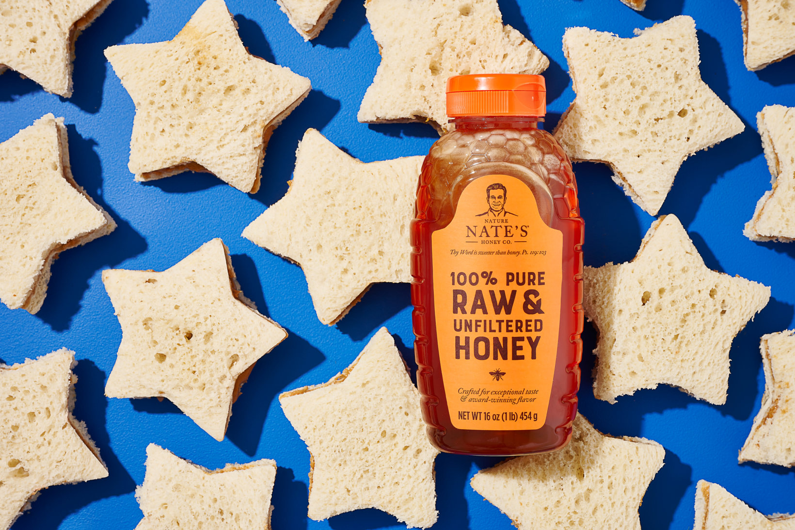 Bottle of Nate's raw and unfiltered honey with an orange cap on blue with star-shaped tea sandwiches around it.