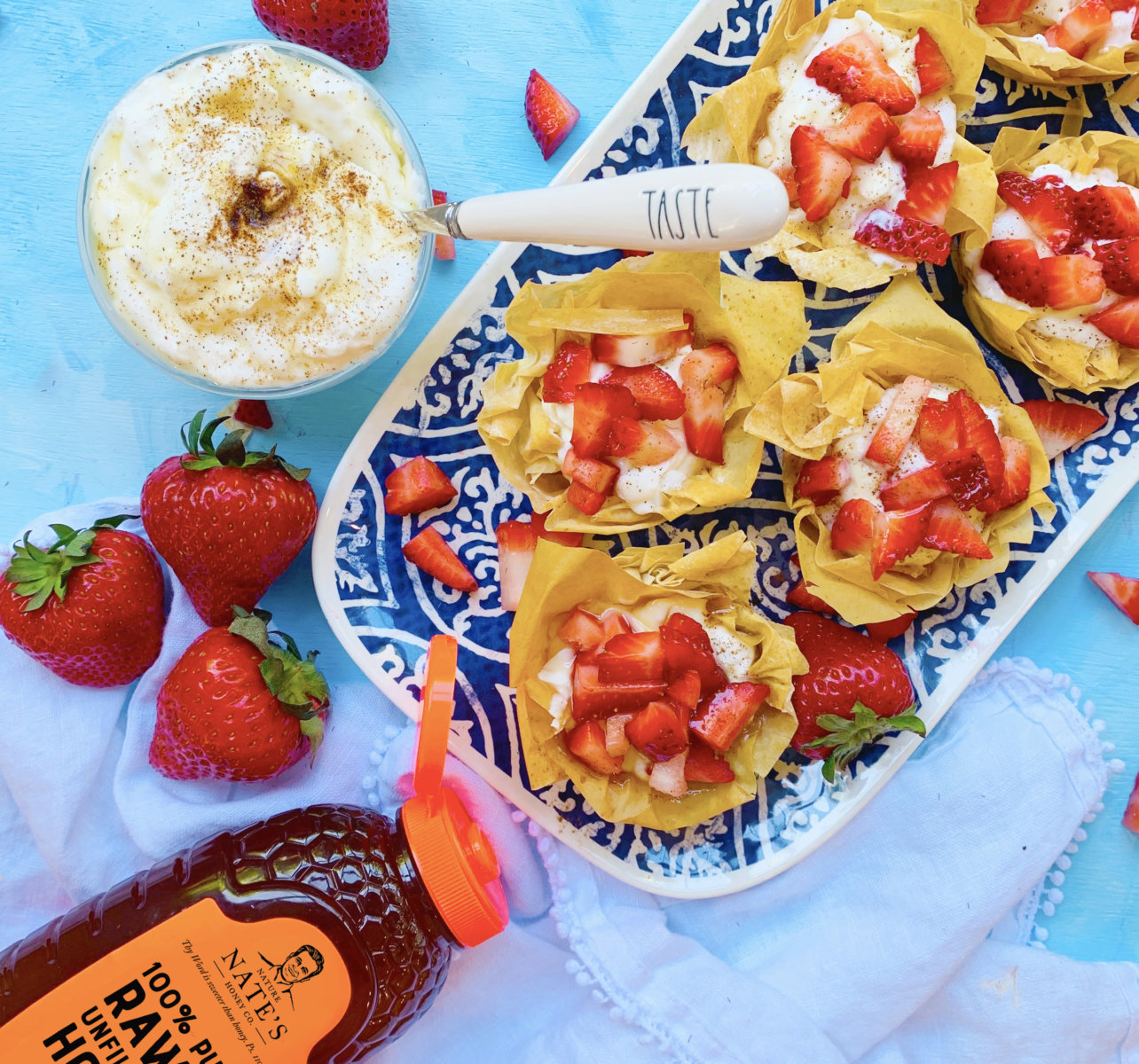 Strawberry filo cups and cream, bowl of yogurt, and Nature Nate's honey bottle on a vibrant blue backdrop.