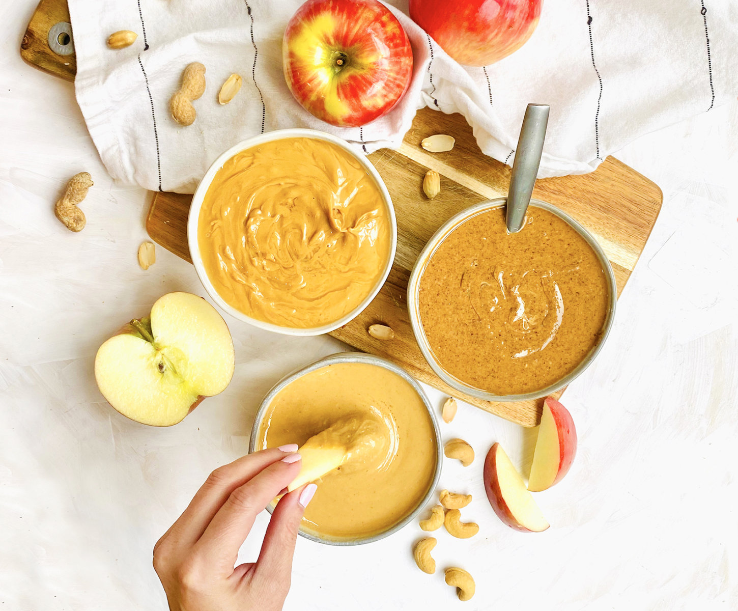 A hand dipping an apple slice into a bowl of honey nut butter with apples and nuts around.