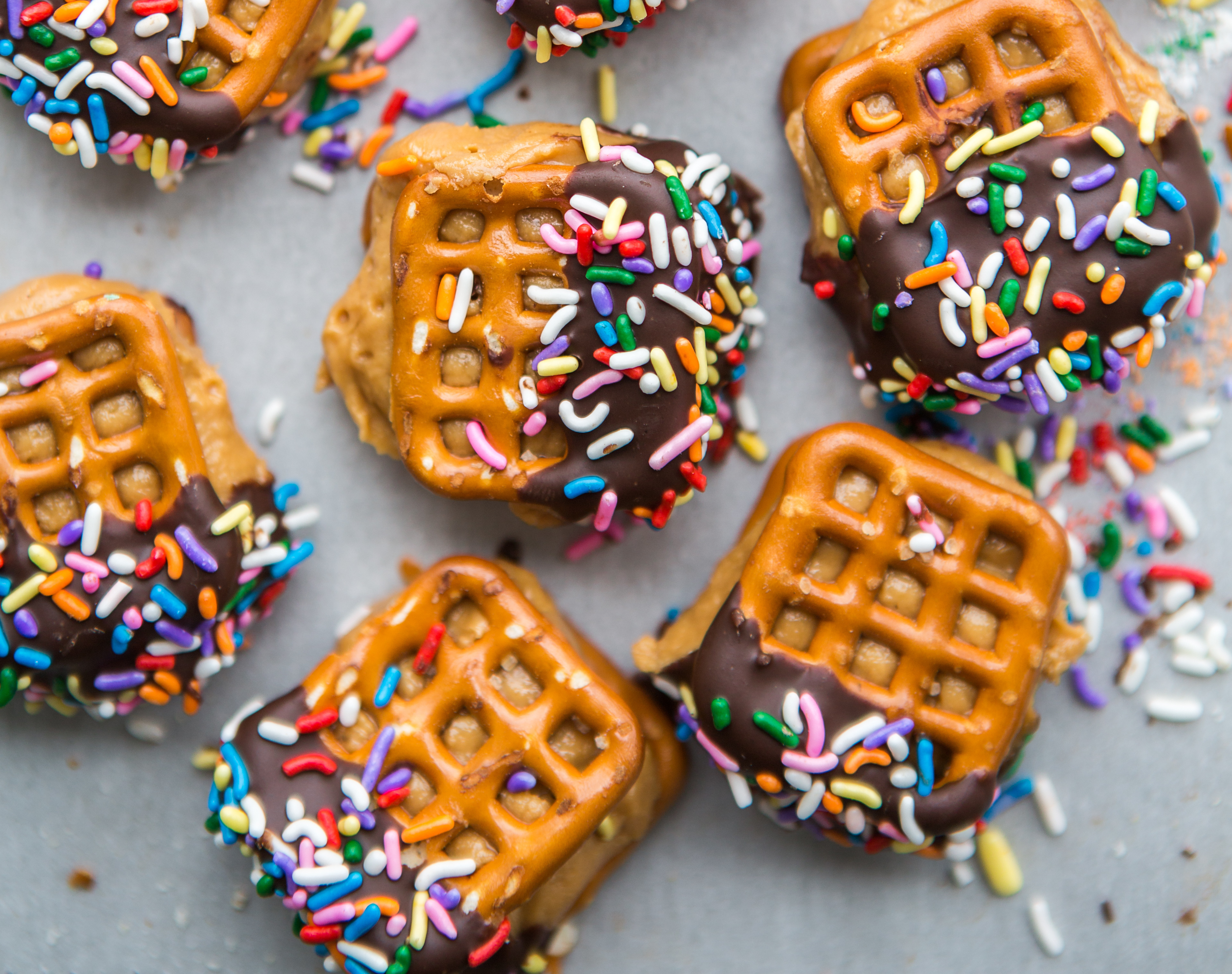 Square pretzels filled with peanut butter and dipped in chocolate and colorful sprinkles on a grey surface.