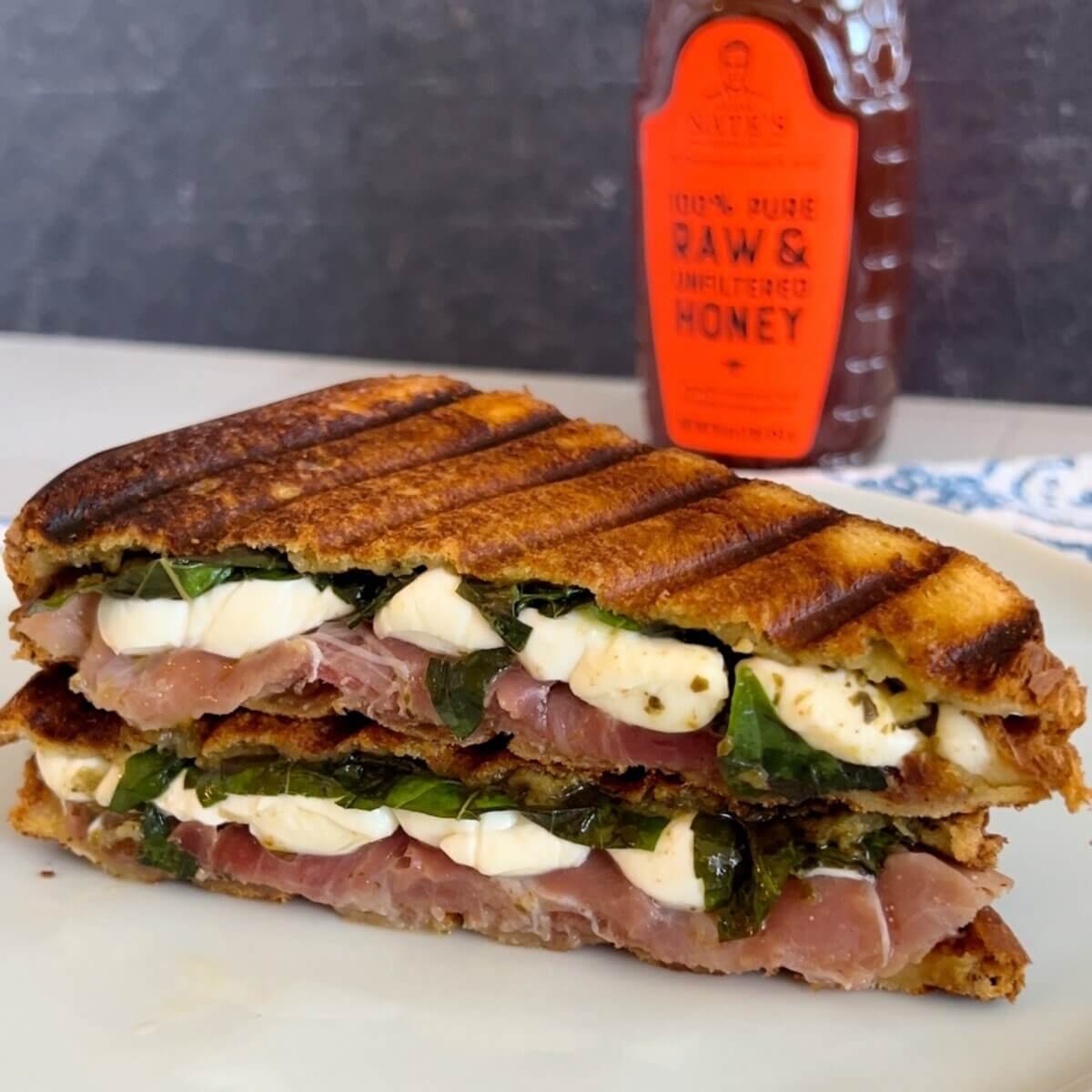 Grilled panini sandwich with basil, prosciutto and mozzarella cheese, next to a bottle of Nate’s honey.