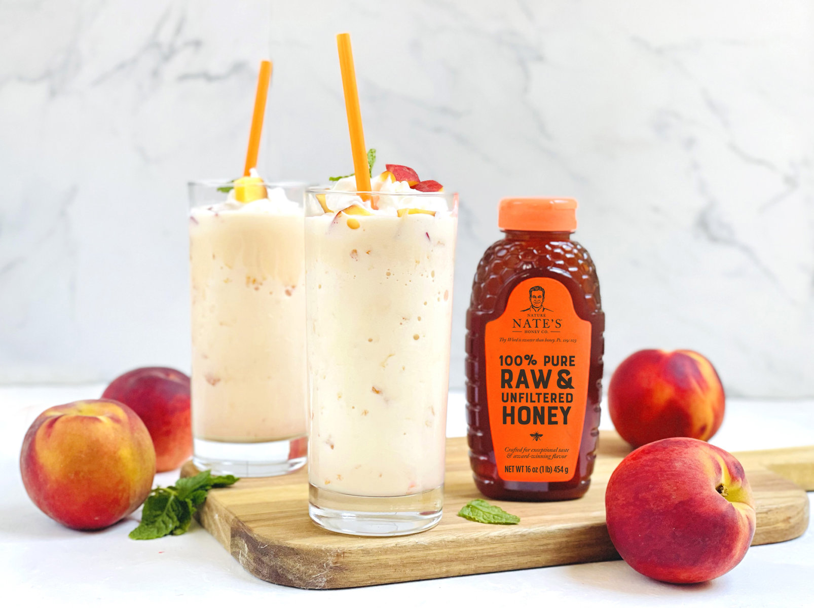 Peach shakes garnished with fruit, with a Nate’s honey bottle and fresh peaches on a wooden board.