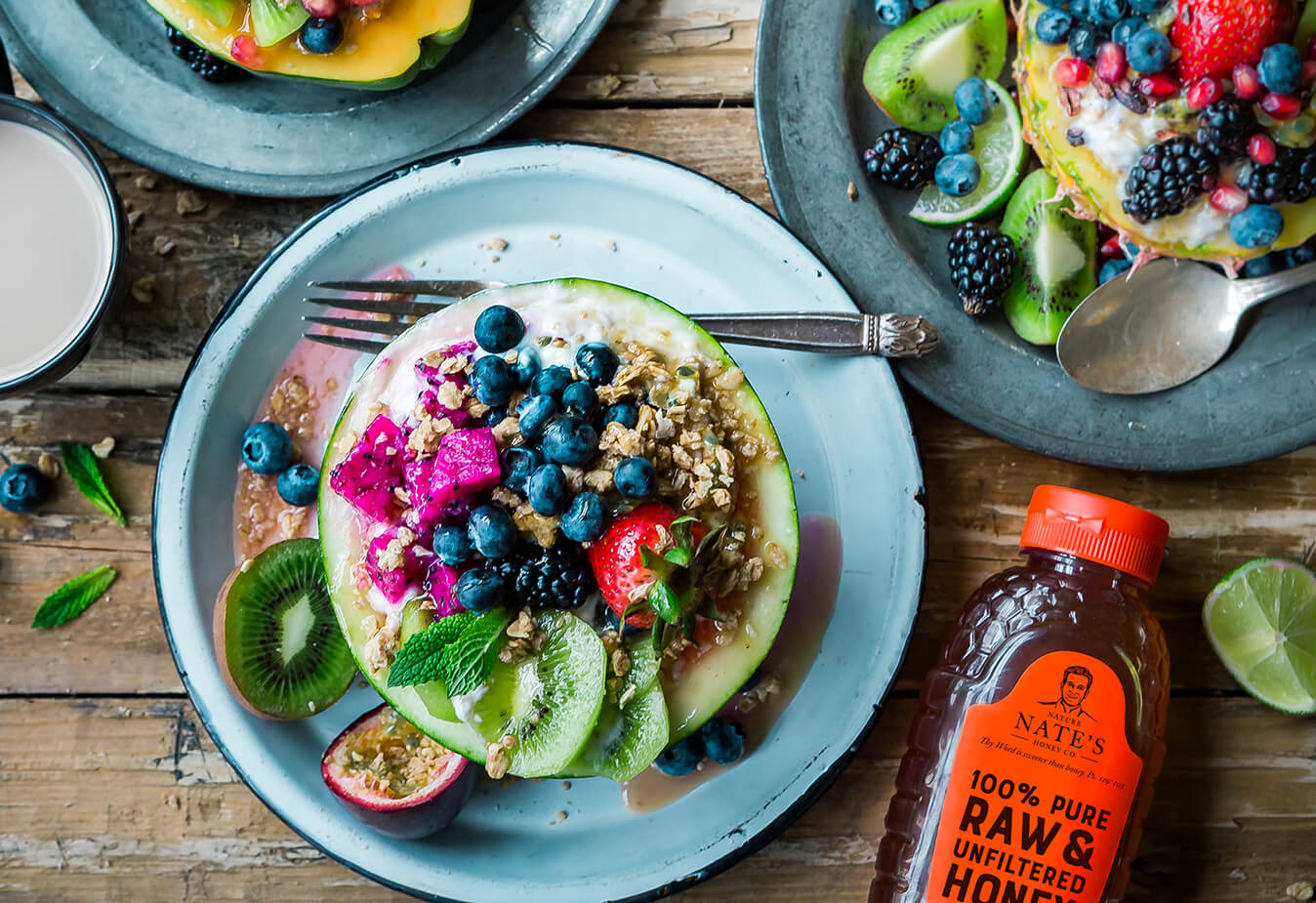 Breakfast bowls with fresh fruits, granola, and yogurt. Nature Nate's 100% Pure Raw & Unfiltered Honey bottle nearby.