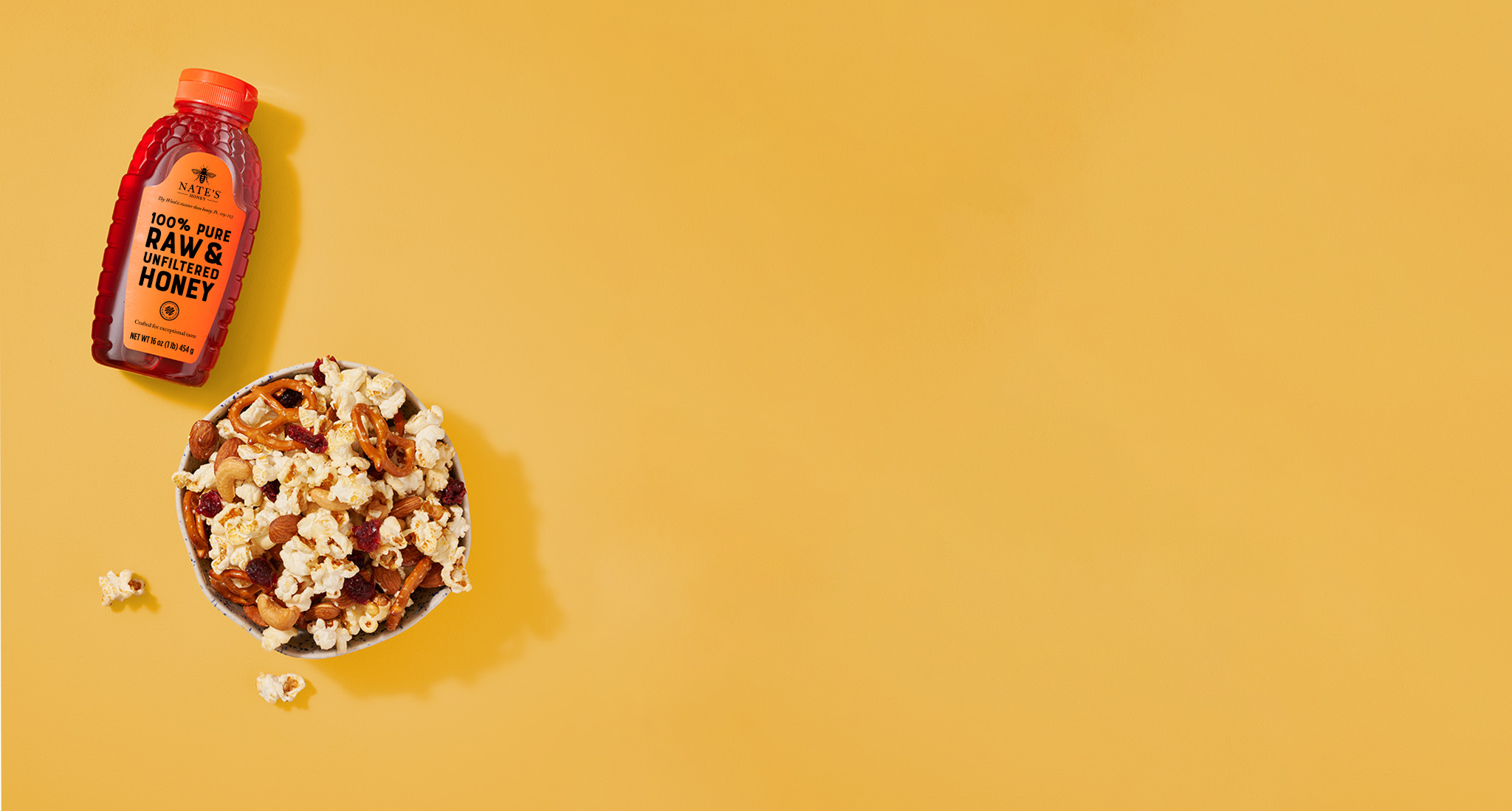 Nate's bottle of honey next to a bowl of popcorn, pretzels and more snack foods on a yellow background.