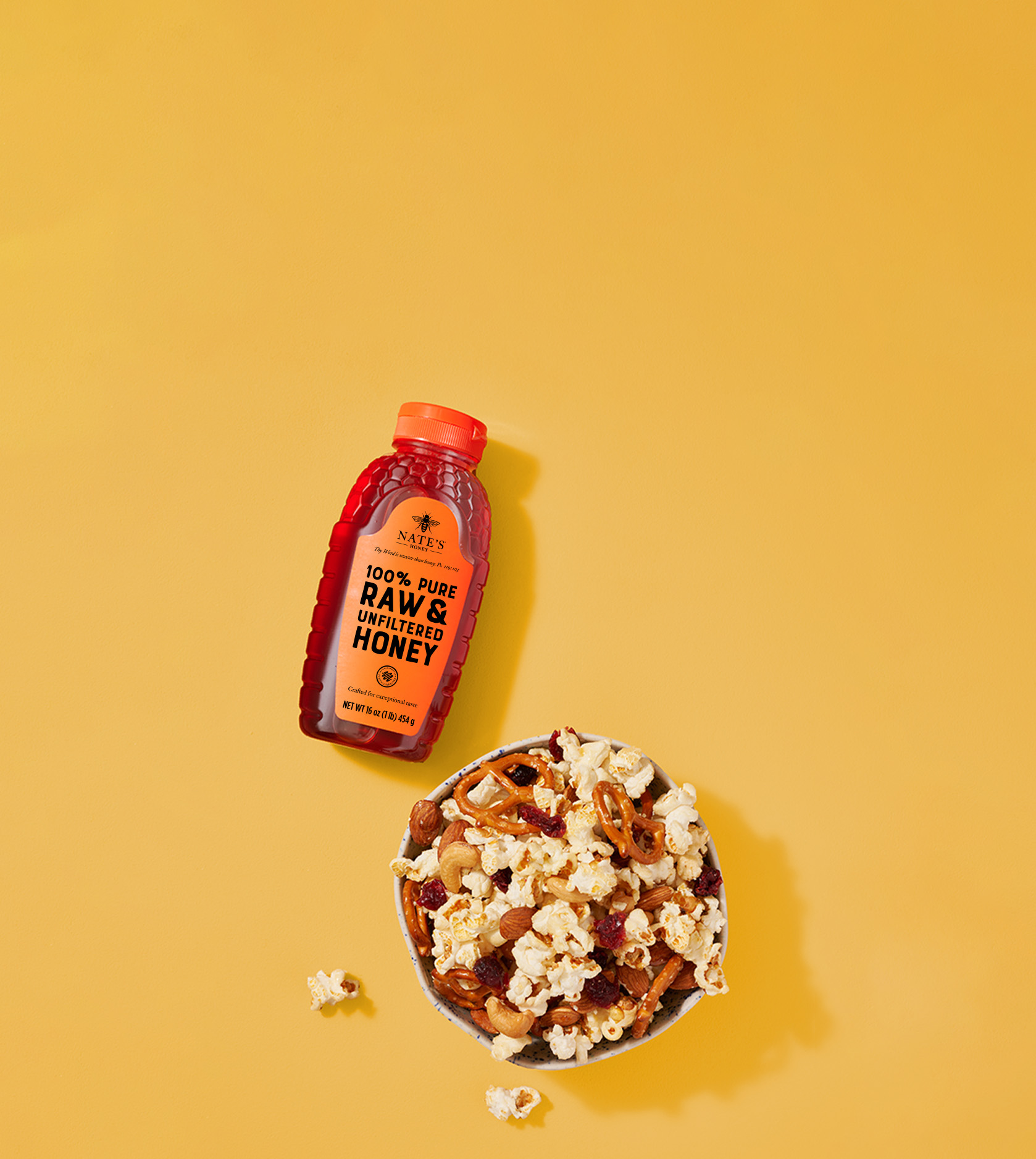 Nate's bottle of honey next to a bowl of popcorn, pretzels and more snack foods on a yellow background.
