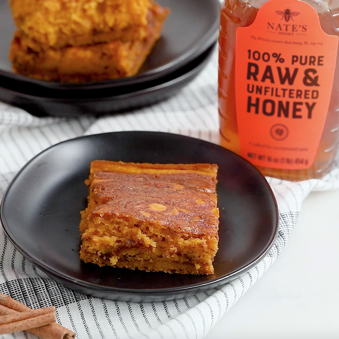 A slice of pumpkin coffee cake on a black plate with a bottle of Nate’s Honey and cinnamon sticks on a checkered cloth.