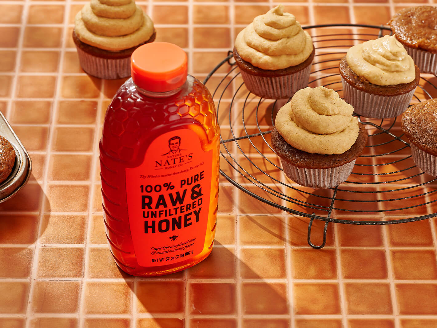Pumpkin cupcakes on a cooling rack and a bottle of Nate’s raw & unfiltered honey on a tiled orange surface.