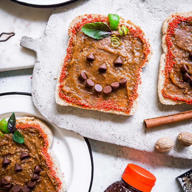 Toast with pumpkin almond spread and smiling face design on a marble board with spices and a Nate's honey bottle nearby.