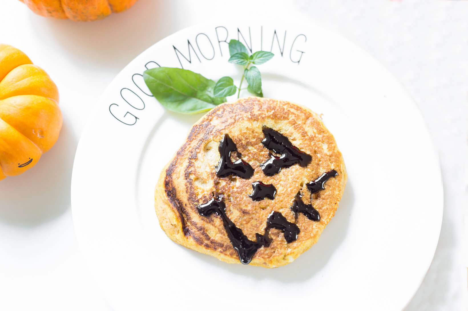 Pumpkin pancake with a smiling face made of chocolate on a white plate with Good Morning text.
