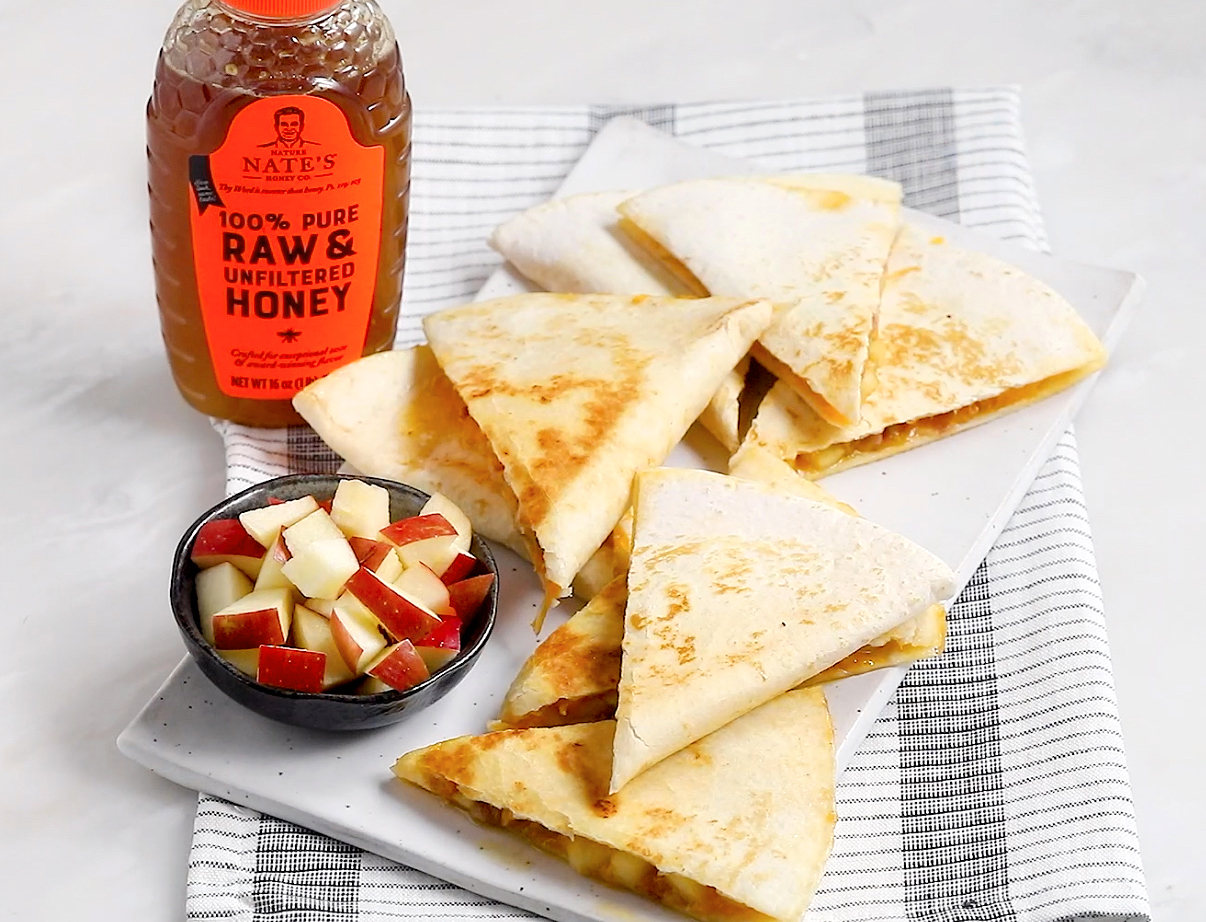 Apple pancetta quesadillas on a plate with diced apples and a bottle of Nate’s raw & unfiltered honey on a striped cloth.