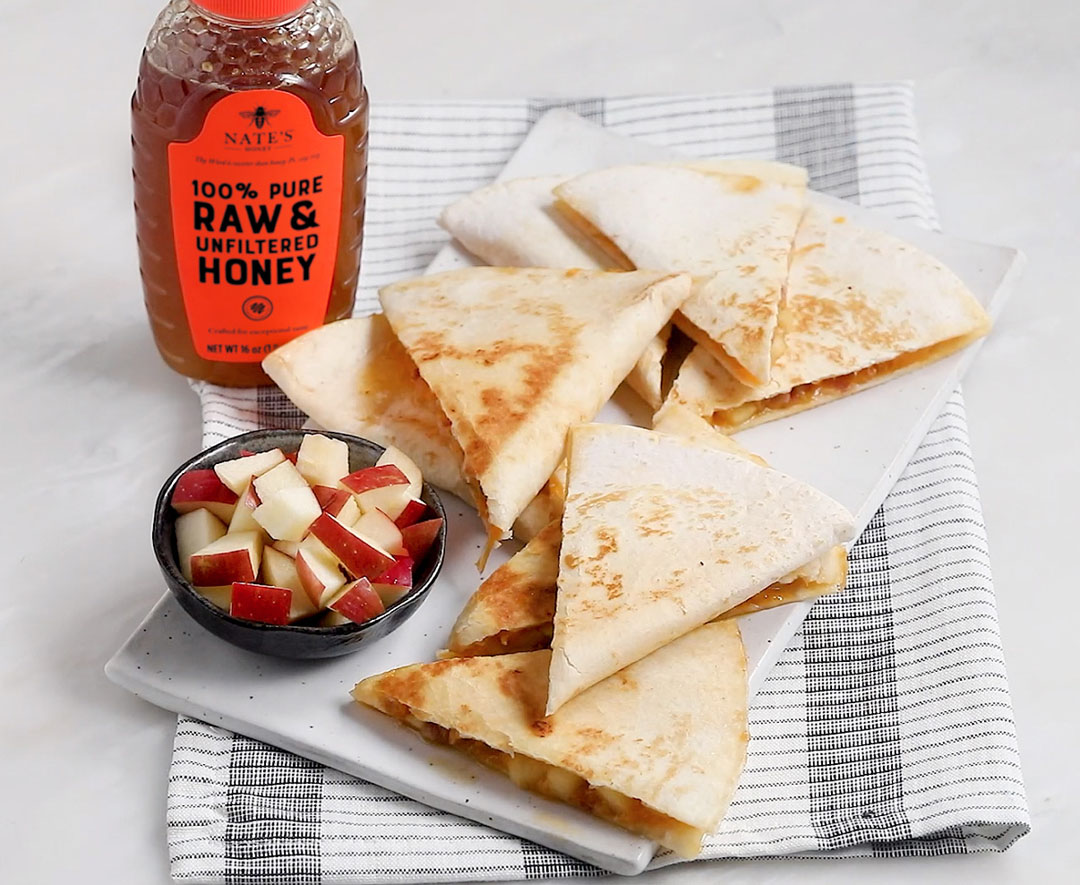Apple pancetta quesadillas on a plate with diced apples and a bottle of Nate’s Honey on a striped cloth.