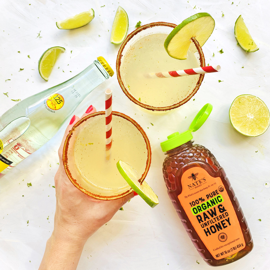 Hand holding a margarita, bottle of soda, bottle of organic Nate’s Honey and lime wedges on white surface.