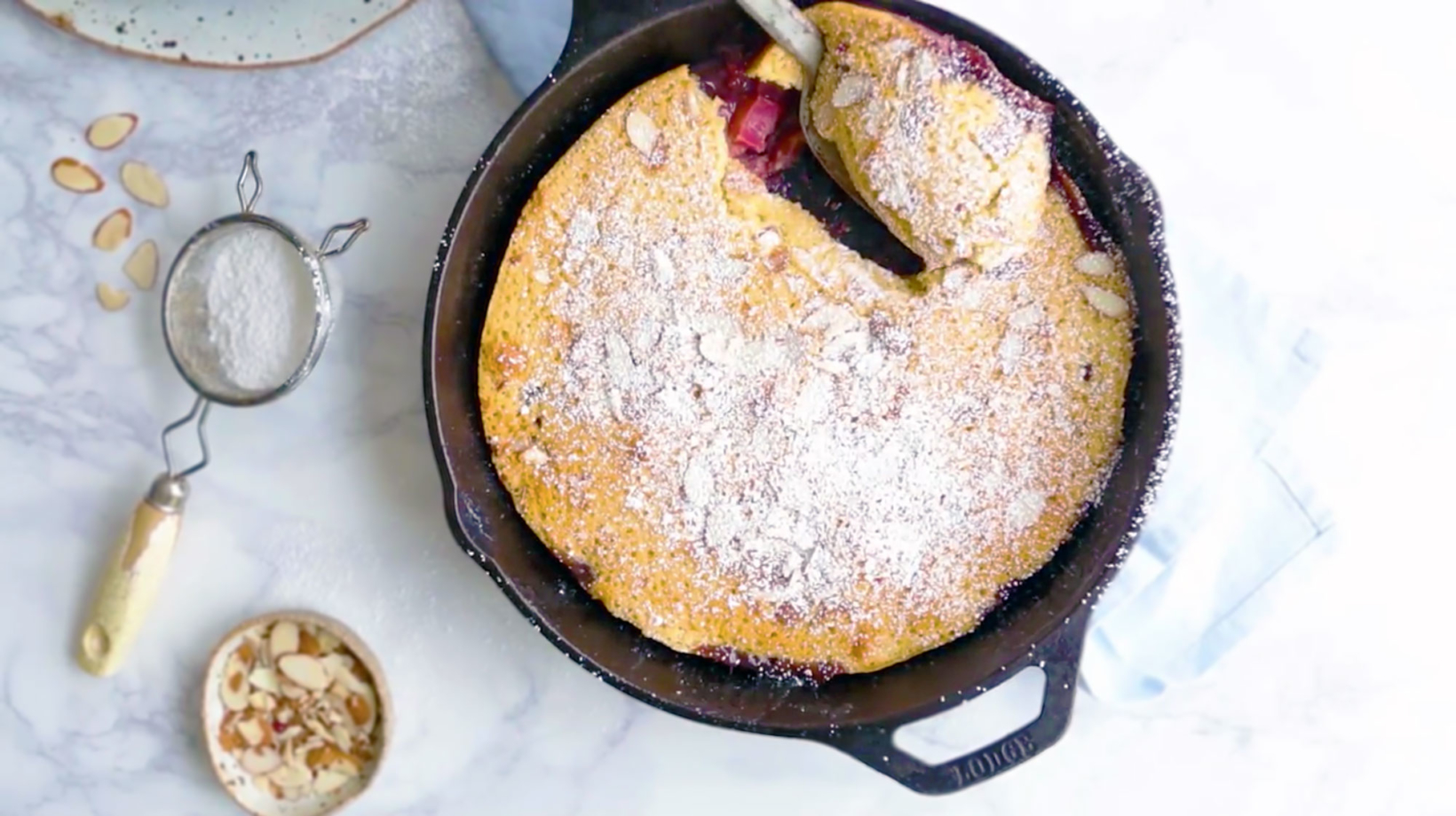 Top view of a rustic berry and rhubarb skillet cake with powdered sugar, almond slivers on side, on a marbled surface.