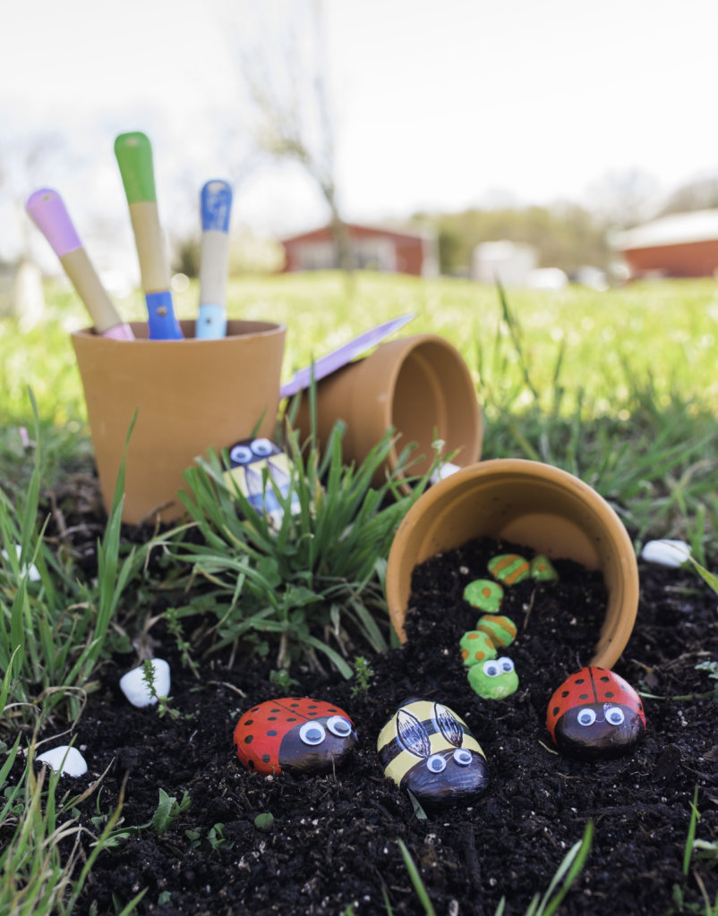 Painted rock insects in soil with pots and garden tags, outdoor craft activity for kids.