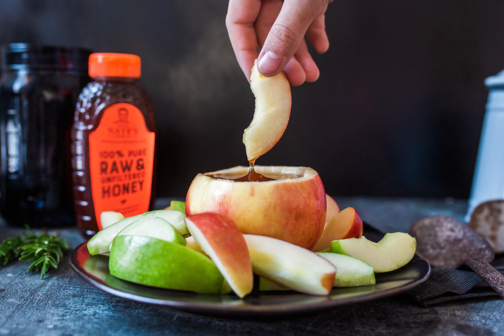 Sliced apples with honey-filled apple, hand dipping slice into honey, two honey bottles in background.