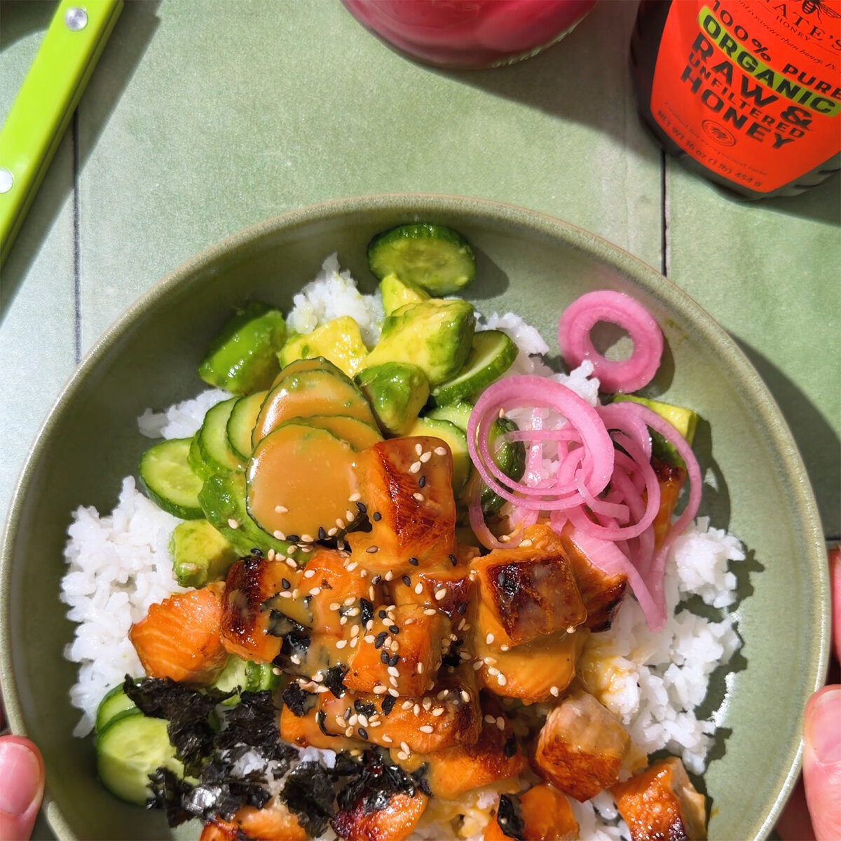 Honey glazed miso salmon bowl with rice, avocado, cucumber salad, pickled onions, and sesame seeds.