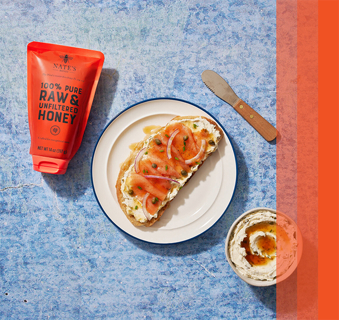 Smoked salmon and cream cheese toast drizzled with honey, next to Nate's honey pouch, knife and bowl on a blue surface.