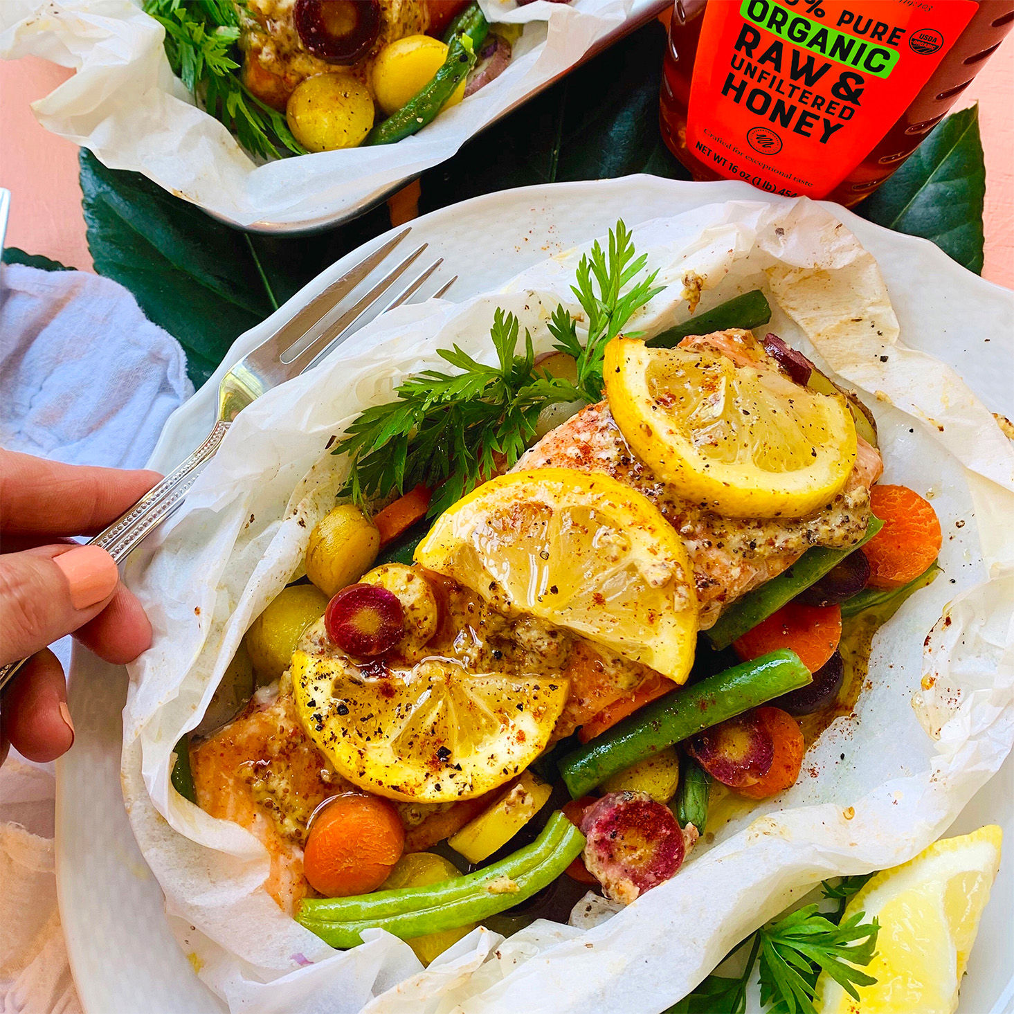 Hand holding fork next to baked salmon packet with veggies and bottle of Nate's Organic Honey.