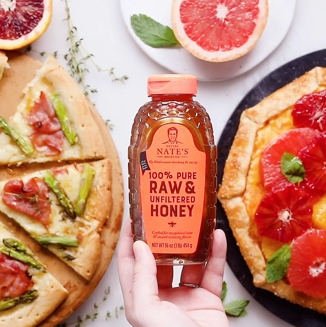 A hand is holding a bottle of Nate's honey in front two pizzas one topped with asparagus and one topped with fruit.