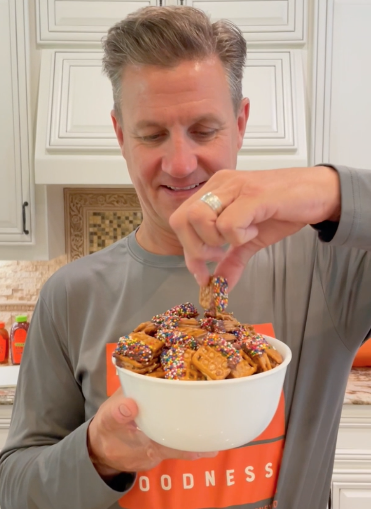 The man Nature Nate smiling choosing a honey pretzel bite from a bowl in a kitchen wearing a 100% PURE GOODNESS shirt.