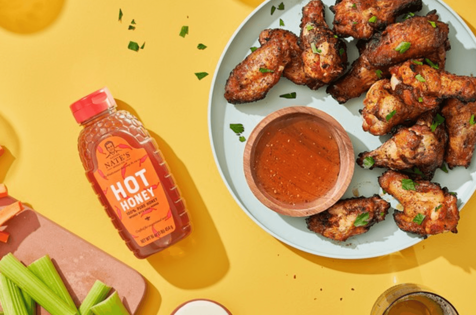 A plate of crispy chicken wings with a bottle of Nate’s hot honey, celery and sauce on a yellow background.