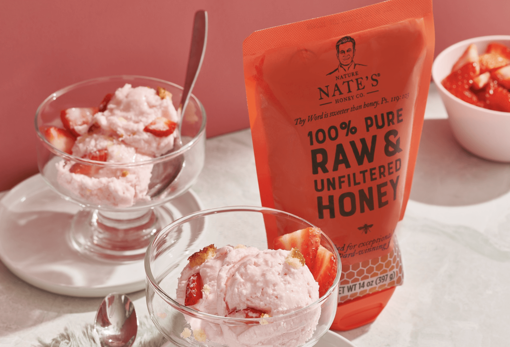 Strawberry ice cream in clear glass bowls drizzled with honey, next to a pouch of Nate’s honey.