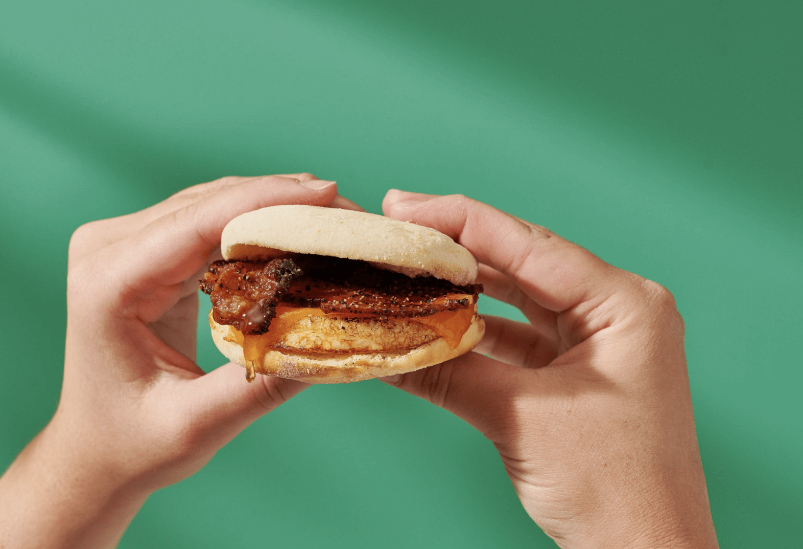 Hands holding a bacon, egg and cheese breakfast sandwich with a drizzle of Nate’s honey against a green background.
