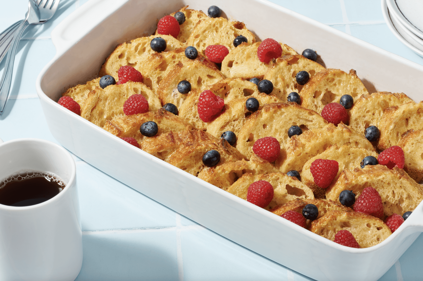 A baking dish of honey vanilla French toast casserole topped with berries, with coffee and cutlery beside it.